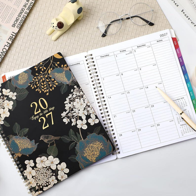 May include: A 2027 spiral-bound planner with a black cover featuring gold and blue floral patterns, a small bird, and the text 2027 Enjoy the little things. The open planner displays a monthly calendar grid with columns for Thursday, Friday, and Saturday.