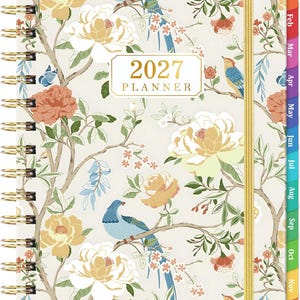 May include: A 2027 planner with a cream-colored cover featuring a botanical pattern of blue birds, orange and yellow flowers, and brown branches. The planner has a gold spiral binding, a gold elastic closure, and colorful monthly index tabs.