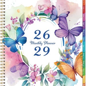 May include: A spiral-bound monthly planner with a white cover featuring a watercolor design of purple, blue, and orange butterflies surrounded by pink and purple flowers. The text 26 Monthly Planner 29 is printed in blue in the center.