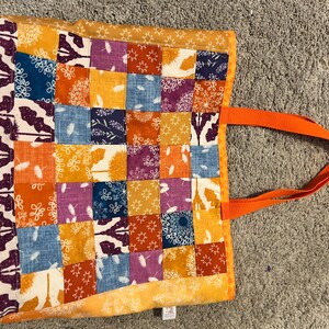 May include: A tote bag featuring a patchwork design of square fabric panels in shades of orange, blue, purple, and white. The bag includes a vertical side panel with a dark purple botanical print and two solid orange fabric handles attached to the side.