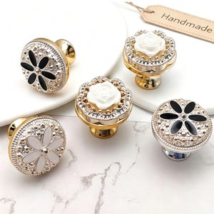 May include: Five round cabinet knobs with metallic gold bases. Two knobs feature black floral petals, one features white floral petals, and two feature raised white rose centers. All knobs have textured, beaded metallic silver rims.