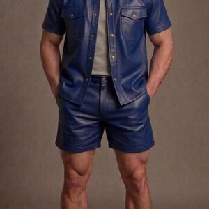 Può includere: Una camicia a maniche corte in pelle blu con due tasche sul petto e pantaloncini in pelle blu abbinati. La camicia è indossata aperta sopra una t-shirt grigia a tinta unita. L'outfit è completato da calzini bianchi e scarpe da ginnastica nere.