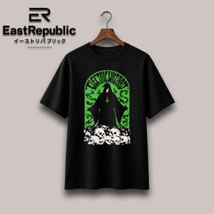 Può includere: Una t-shirt nera a maniche corte con una stampa grafica di una figura incappucciata in tunica con una croce rovesciata, circondata da un arco verde e fumo, con un mucchio di teschi bianchi alla base e il testo Electric Wizard sopra.