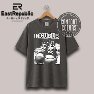 Puede incluir: Una camiseta de manga corta gris carbón con un estampado gráfico blanco de un par de zapatillas altas. El texto INCUBUS está impreso sobre las zapatillas. La camiseta tiene cuello redondo y se muestra en una percha de madera con etiqueta.