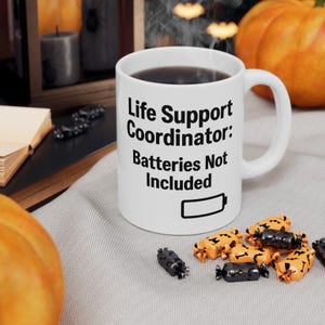 May include: A white ceramic mug featuring black text which reads Life Support Coordinator: Batteries Not Included, alongside a graphic of an empty battery icon. The mug has a standard handle and is filled with a dark liquid, likely coffee or tea.