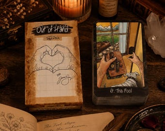 Puede incluir: Una caja de baraja de tarot titulada Out of Hand con un dibujo lineal de manos formando un corazón, y una carta de tarot titulada 0. The Fool que muestra a una persona con un perro mirando por una ventana. La caja tiene un patrón de cuadrícula.