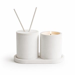 May include: A white cylindrical concrete diffuser with two sticks and a matching white cylindrical concrete candle with a lit wick, both resting on an oval white concrete tray. The set features a smooth, matte finish and a uniform light color.