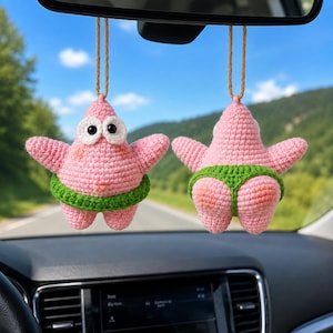 Pode incluir: Dois ornamentos de estrela-do-mar em crochê rosa pendurados por cordões. A figura da esquerda mostra uma vista frontal com grandes olhos brancos e uma faixa verde. A figura da direita mostra uma vista traseira com uma peça verde tipo fio-dental.