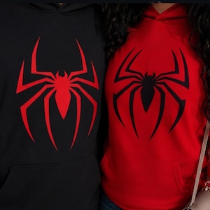 May include: Two hooded sweatshirts featuring a large spider graphic on the chest. One hoodie is black with a red spider emblem, while the other is red with a black spider emblem. Both garments include a front kangaroo pocket and a hood.