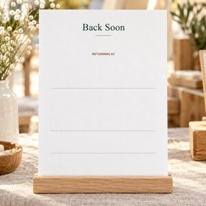 Op de afbeelding: Een wit rechthoekig bord in een standaard van licht hout. Het bord bevat de donkergroene tekst Back Soon, een dunne horizontale lijn en de oranje tekst RETURNING AT boven drie lege horizontale lijnen voor informatie.
