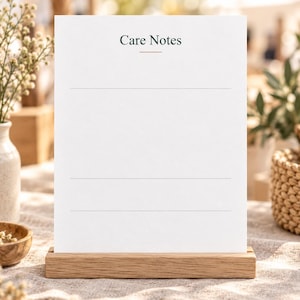Op de afbeelding: Een witte rechthoekige kaart met de tekst Care Notes in donkergroen bovenaan, voorzien van een dunne horizontale lijn onder de tekst en drie extra dunne horizontale lijnen eronder, geplaatst in een lichtbruine houten standaard.