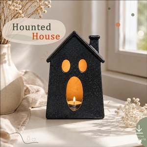 May include: A black house-shaped candle holder with a textured finish, featuring two oval eye cutouts and a large oval door cutout. A lit tea light candle sits inside the structure, emitting a warm glow. The text "Haunted House" is visible above.