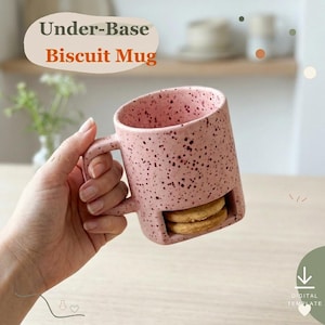 May include: A pink ceramic mug with dark speckles featuring a built-in horizontal slot at the base for storing biscuits. The text Under-Base Biscuit Mug is printed at the top. The mug has a sturdy handle and holds two round light brown cookies.