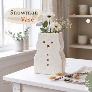 Op de afbeelding: Een gespikkelde gebroken witte keramische vaas in de vorm van een sneeuwman met twee ronde ogen, een driehoekige neus en drie ronde knopen. De vaas bevat witte madeliefjes en groene eucalyptusbladeren. De tekst Snowman Vase is zichtbaar.