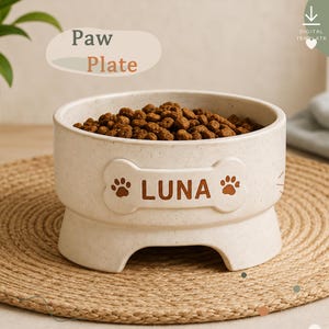 Puede incluir: Un cuenco para mascotas de color blanco roto moteado con un emblema en relieve en forma de hueso en la parte frontal. El emblema presenta el nombre LUNA en letras mayúsculas marrones, flanqueado por dos huellas de pata marrones. Lleno de pienso.