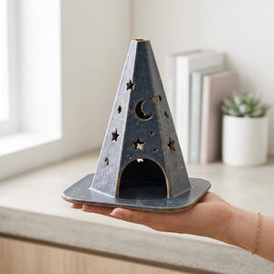 May include: A cone-shaped ceramic incense burner in a dark blue speckled finish. The structure features cutouts of stars and a crescent moon, with a wide arched opening at the base and a flat square-edged platform for stability and placement.