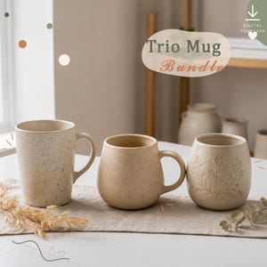 May include: A trio of ceramic mugs in neutral beige tones. The first mug is tall with dark speckles, the second is a rounded smooth mug, and the third features an embossed leaf pattern. Text at the top reads Trio Mug Bundle.
