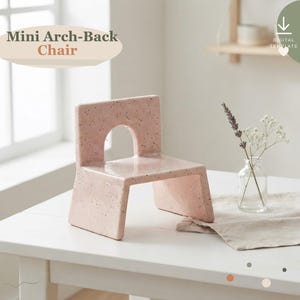 May include: A light pink mini chair with a speckled finish and an arched cutout in the backrest. The text "Mini Arch-Back Chair" is visible in the upper left corner. The chair has a smooth, glossy surface and a wide, stable base.