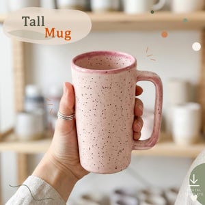 May include: A tall ceramic mug with a light pink speckled finish and a darker pink rim. The mug features a rectangular handle and the text "Tall Mug" is printed in the upper left corner. The design has a smooth, glazed texture throughout.