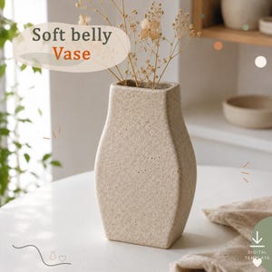 Op de afbeelding: Een beige, getextureerde vaas met een zachte buikvorm, gevuld met gedroogde bloemen. De tekst "Soft belly Vase" is zichtbaar in de linkerbovenhoek. De vaas heeft een gespikkelde, matte afwerking en een rechthoekige basis die naar boven toe smaller wordt.