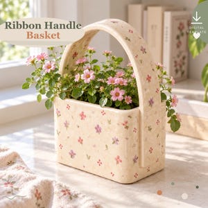 May include: A cream-colored ceramic basket with a high arched handle, decorated with a small pink and purple floral pattern. The basket is filled with a green plant featuring small pink flowers. Text reads Ribbon Handle Basket.