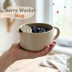 May include: A speckled beige ceramic mug with a built-in strainer section near the rim, filled with fresh blueberries. The text Berry Washer Mug is printed above the mug. The design includes a handle on the side and a wide, open top.