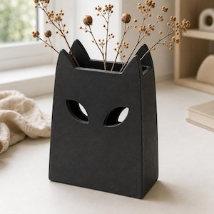 Könnte beinhalten: Eine mattschwarze Keramikvase in Form eines Katzenkopfes mit spitzen Ohren und zwei mandelförmigen Aussparungen als Augen. Die Vase enthält mehrere dünne, braune, getrocknete botanische Stängel mit kleinen runden Samenkapseln.