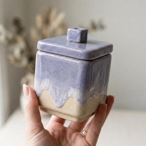 May include: A square ceramic canister with a lid featuring a small square knob. The container has a two-tone finish with a light beige base and a lavender glaze that drips down from the top, creating a textured, layered appearance on the sides.