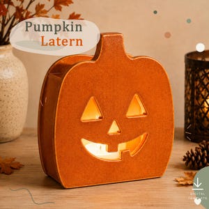 Może przedstawiać: Płaska, pomarańczowa latarnia w kształcie dyni z wyciętą twarzą jack-o'-lantern, posiadająca dwoje trójkątnych oczu, trójkątny nos i szeroki, wyszczerbiony uśmiech. Latarnia jest podświetlona od wewnątrz, emitując ciepły blask przez wycięcia.