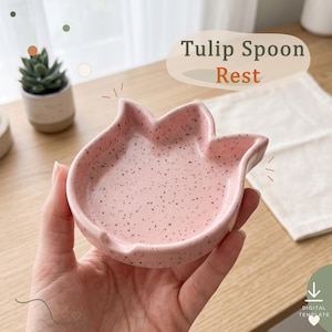 Może przedstawiać: Jasnoróżowa ceramiczna podstawka pod łyżkę w kształcie kwiatu tulipana z nakrapianym wykończeniem. Powierzchnia posiada drobne ciemnobrązowe lub czarne kropki rozmieszczone w miseczce. W prawym górnym rogu widoczny jest napis Tulip Spoon Rest.