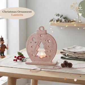 May include: A dusty pink ceramic Christmas ornament lantern featuring a cutout pine tree shape in the centre, surrounded by small star and circle perforations. A small white tea light candle sits inside the base of the ornament.