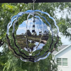 May include: A circular metal wind spinner featuring a printed image of a black and white striped lighthouse standing near a marshy landscape with a wooden walkway. The spinner has multiple concentric scalloped layers that create a 3D effect.