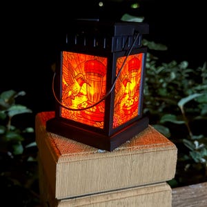 May include: A square black lantern with a glowing red stained glass effect featuring a hummingbird and a feeder design. The lantern has a dark frame, a top handle, and emits a warm light from within, showcasing detailed illustrations on its panels.