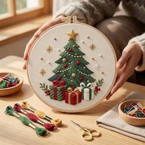 May include: A circular wooden embroidery hoop containing a fabric panel with a hand-stitched Christmas tree design. The tree is decorated with red and gold ornaments, a gold star topper, and sits above three gift boxes in red, green, and white.