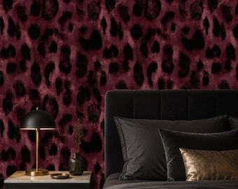 Op de afbeelding: Een slaapkamer met een donkergrijs gestoffeerd hoofdeinde, donkergrijze kussens en een goudkleurig sierkussen. Een zwarte koepelvormige lamp staat op een zwart nachtkastje tegen een muur met een donkerrood en zwart luipaardpatroon.