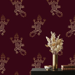 May include: A deep burgundy wallpaper featuring a repeating pattern of gold-colored line art geckos. In the foreground, a tall metallic gold vase holds a bouquet of dried cream and gold-toned foliage next to a small bowl and two stacked books.