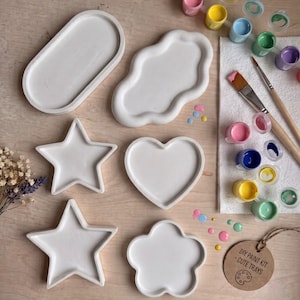 Può includere: Un kit di pittura fai-da-te con sei vassoi bianchi a forma di ovale, nuvola, stella, cuore e fiore, insieme a due pennelli, un set di piccoli barattoli di vernice di vari colori e un'etichetta rotonda marrone con scritto DIY PAINT KIT - CUTE TRAYS.