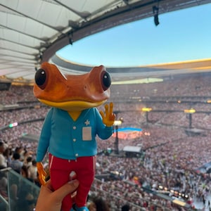 Puede incluir: Una figura de rana con una cabeza grande de color naranja y ojos negros, que viste una camisa azul claro abotonada con un pequeño emblema de estrella blanca y pantalones rojos. La rana tiene piel amarilla en el cuello y el pecho.