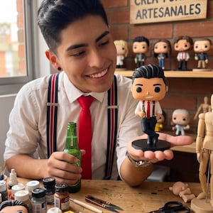 Könnte beinhalten: Eine individuelle Vinyl-Sammelfigur, die eine Figur mit dunklem Haar, weißem Hemd, roter Krawatte, dunkelblauer Hose und Hosenträgern darstellt, die eine kleine grüne Flasche hält und auf einem runden schwarzen Sockel steht.