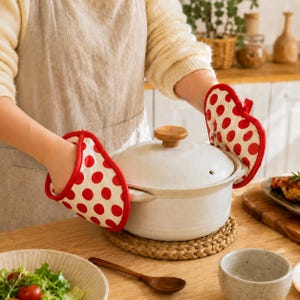 Peut inclure: Une paire de gants de cuisine blancs avec un motif à pois rouges et une bordure rouge. Les gants sont conçus pour couvrir les mains et servent à tenir une marmite en céramique blanche avec un couvercle assorti et un bouton en bois.