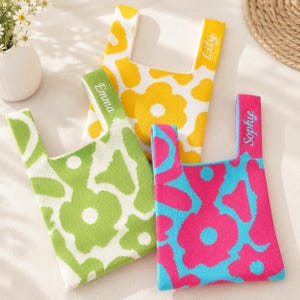 May include: Three knit tote bags featuring floral patterns in yellow and white, green and white, and pink and blue. Each bag handle is personalised with embroidered names: Lily, Emma, and Sophie. The bags have a flat, rectangular shape.