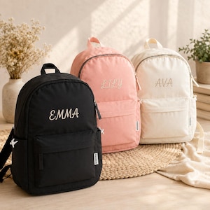 Peut inclure: Trois sacs à dos en noir, rose clair et crème, chacun doté d'une poche avant, d'une poignée supérieure et d'un nom brodé en écriture cursive sur le devant. Le sac noir porte le nom Emma, le rose Lily et le crème Ava.
