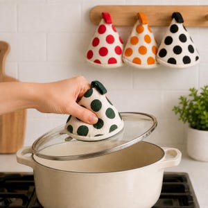May include: A cone-shaped pot holder with a cream base and dark green polka dots, held over a glass lid on a white pot. Three additional cone-shaped pot holders in red, orange, and black polka dot patterns hang on a wooden rack in the background.