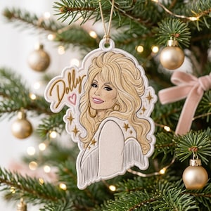 May include: A white, die-cut hanging ornament featuring a detailed illustration of a blonde person with voluminous hair, wearing a fringed garment. The word Dolly is written in gold script next to a small pink heart, accented by gold star icons.