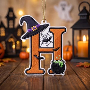 May include: An embroidered hanging ornament in the shape of the letter H, featuring an orange base with a black witch hat, a white spider web, black bats, and a black cauldron filled with bubbling green liquid, suspended by a gold cord.
