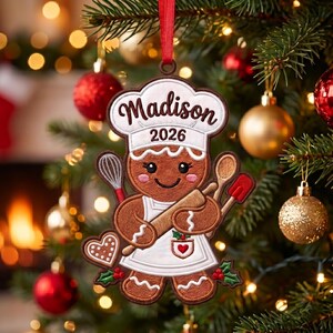 Könnte beinhalten: Ein brauner Lebkuchenmann-Anhänger mit weißer Kochmütze und Schürze, bestickt mit dem Namen Madison und dem Jahr 2026. Die Figur hält ein Nudelholz, einen Schneebesen und einen Teigschaber sowie einen kleinen herzförmigen Keks und Stechpalmen.