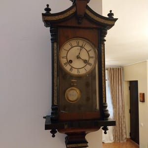 May include: A wall-mounted wooden clock featuring a dark brown finish, a circular white face with black Roman numerals, and ornate decorative accents. The clock includes a glass-fronted lower section revealing a circular brass pendulum.