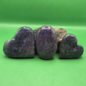 May include: Three polished stones with a dark green and deep red mottled texture. Two stones are heart-shaped, one on the left and one on the right, while the central stone is oval-shaped. The surfaces show natural mineral inclusions.