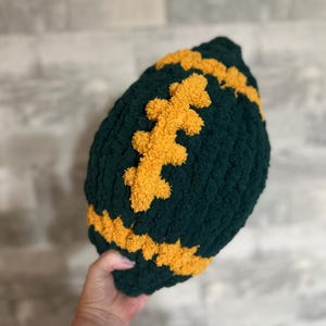 May include: A plush, hand-crocheted football made from dark green yarn with bright yellow yarn accents. The football features a textured, fuzzy surface with a central yellow lace detail and yellow stripes at both ends of the oval-shaped object.