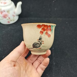 May include: A small, beige ceramic tea cup featuring a hand-painted illustration of a dark gray mouse reaching toward a cluster of red berries with green leaves. The cup has a speckled texture and a smooth, rounded rim, with a matching teapot visible.
